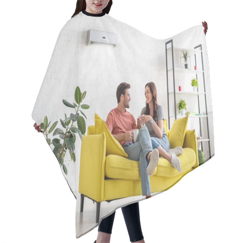 Personality  Happy Young Couple Talking And Holding Cups While Sitting On Yellow Sofa Under Air Conditioner At Home Hair Cutting Cape