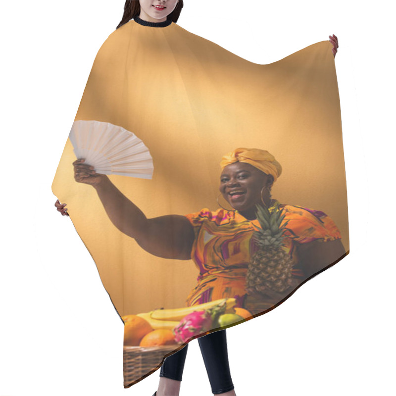 Personality  Smiling Middle Aged African American Woman Holding Pineapple Near Fruits And Waving With Fan On Orange Hair Cutting Cape