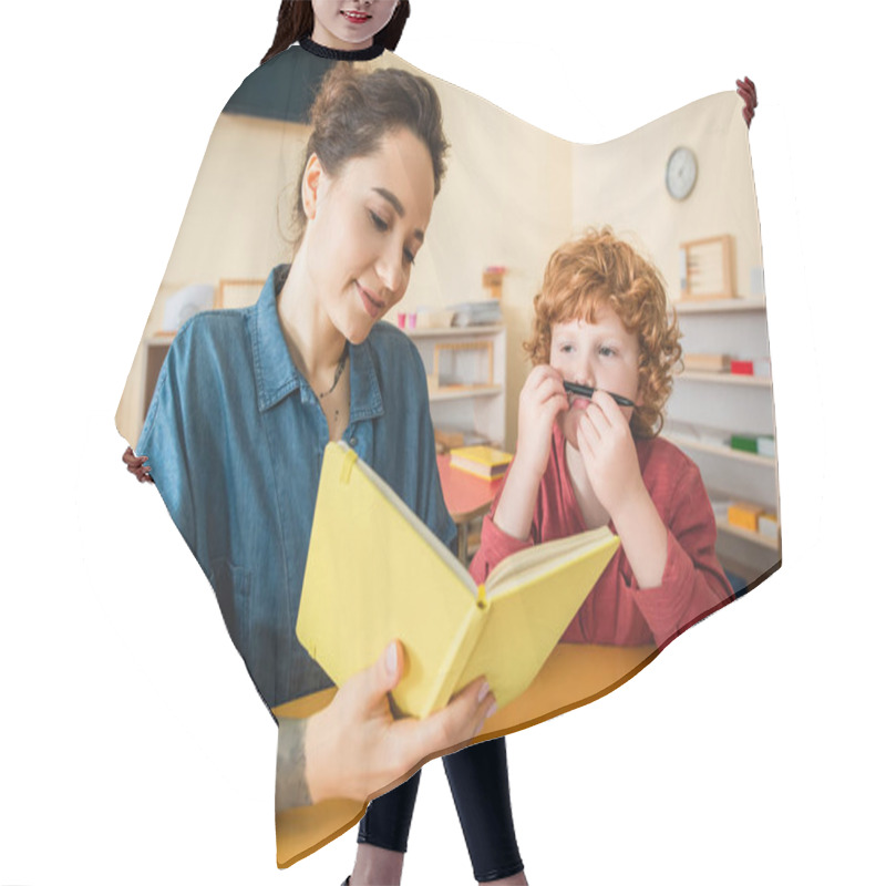 Personality  Boy Holding Pen Near Face While Montessori School Teacher Reading Book Hair Cutting Cape