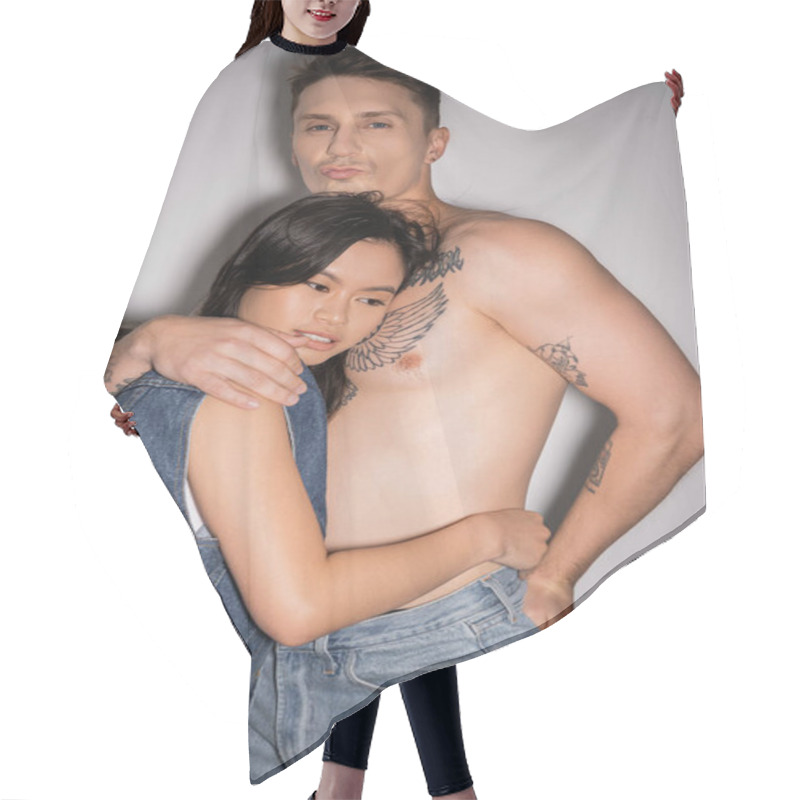 Personality  Tattooed And Muscular Man Pouting Lips And Hugging Asian Woman In Denim Vest On Grey Background Hair Cutting Cape