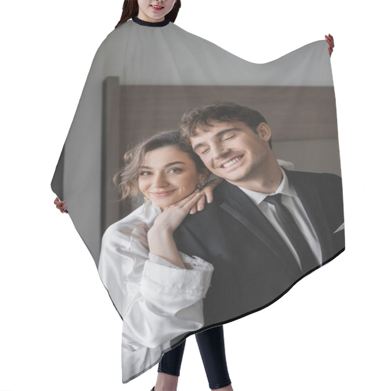 Personality  Happy Bride In Jewelry And White Silk Robe Leaning On Shoulder Of Joyful Groom In Classic Black Suit While Standing Together In Modern Hotel Room During Honeymoon, Newlyweds  Hair Cutting Cape