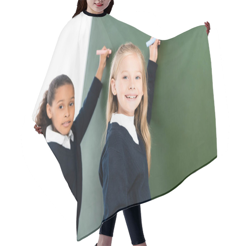 Personality  Two Smiling Multicultural Schoolgirls Writing On Chalkboard While Looking At Camera  Hair Cutting Cape