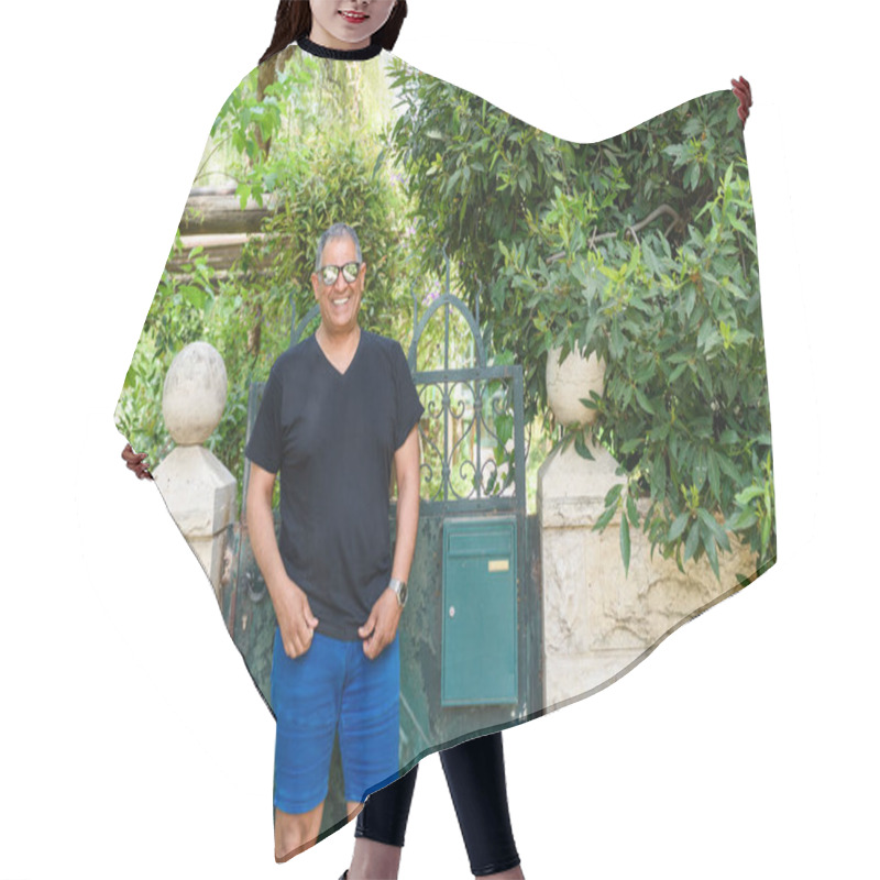 Personality  Joyful Senior Man In Casual Black T-shirt And Blue Shorts Stands By Rustic Green Gate, Surrounded By Lush Greenery In Tranquil Garden Setting.Perfect For Themes Of Travel, Tourism, And Outdoor Leisure Hair Cutting Cape