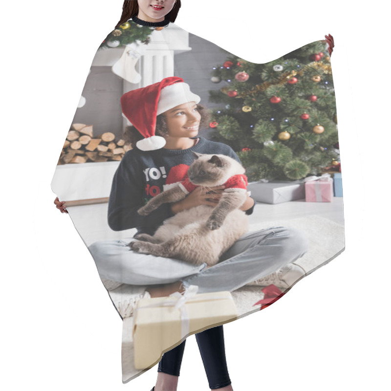 Personality  Happy African American Girl In Santa Hat Looking Away While Sitting Near Christmas Tree And Holding Fluffy Cat Hair Cutting Cape