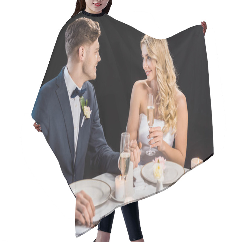 Personality  Happy Young Couple Sitting At Served Table With Glasses Of Champagne And Looking At Each Other Isolated On Black Hair Cutting Cape