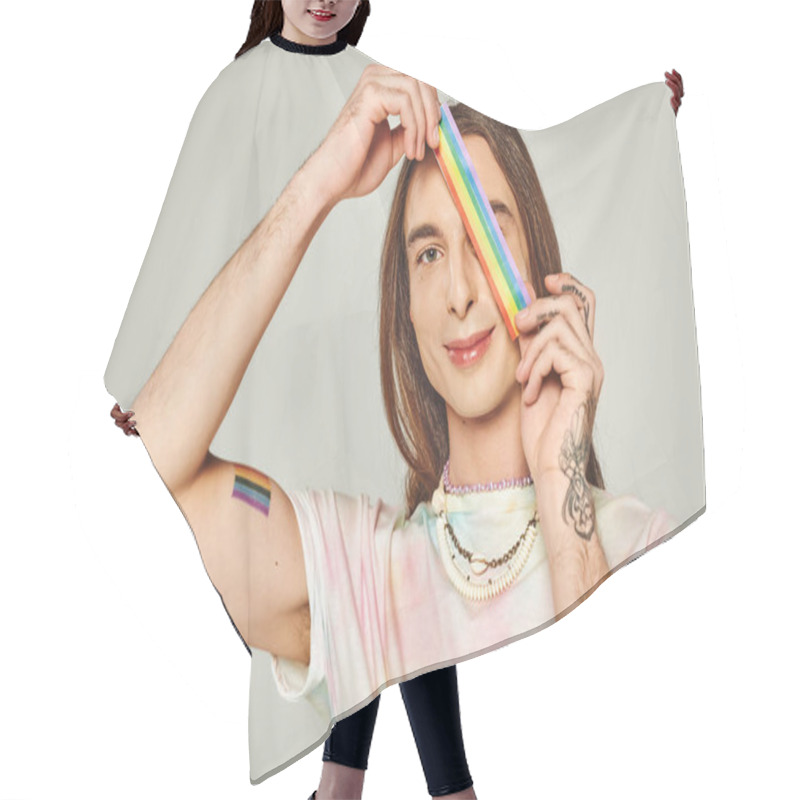 Personality  Happy And Tattooed Gay Man With Long Hair And Tie Dye T-shirt Holding Rainbow Lgbt Flag Near Face During Pride Month And Smiling While Looking At Camera On Grey Background  Hair Cutting Cape