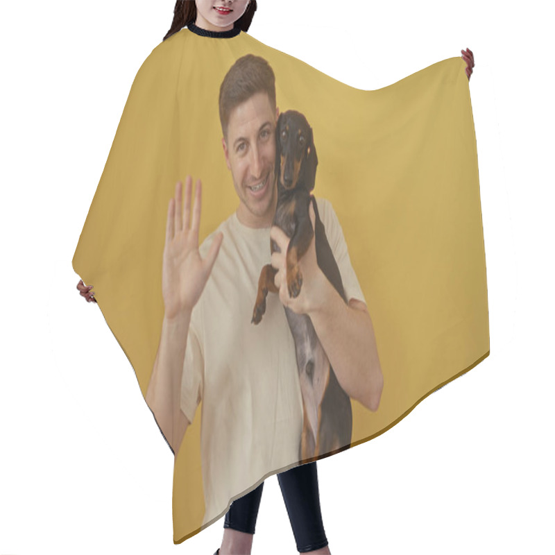 Personality  Hispanic Man Holding His Dachshund Dog While Waving Against A Vibrant Yellow Background, Showcasing A Cheerful And Pet-friendly Moment. Hair Cutting Cape