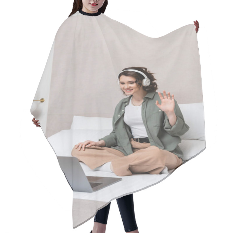 Personality  Overjoyed Woman With Wavy Brunette Hair, In Wireless Headphones And Casual Clothes Sitting On Bed And Waving Hand During Video Call On Laptop Near White Pillows And Wall Sconce In Hotel Room Hair Cutting Cape