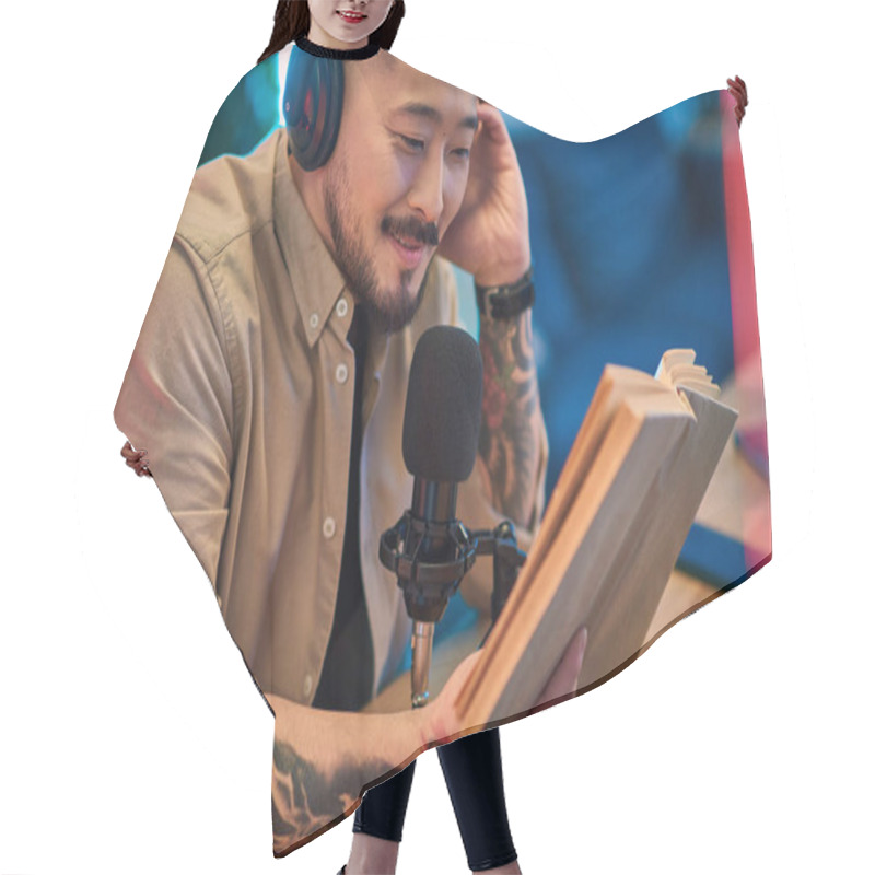 Personality  An Asian Man Sits At A Desk In His Home Studio, Wearing Headphones And Holding A Book While Recording A Podcast. Hair Cutting Cape