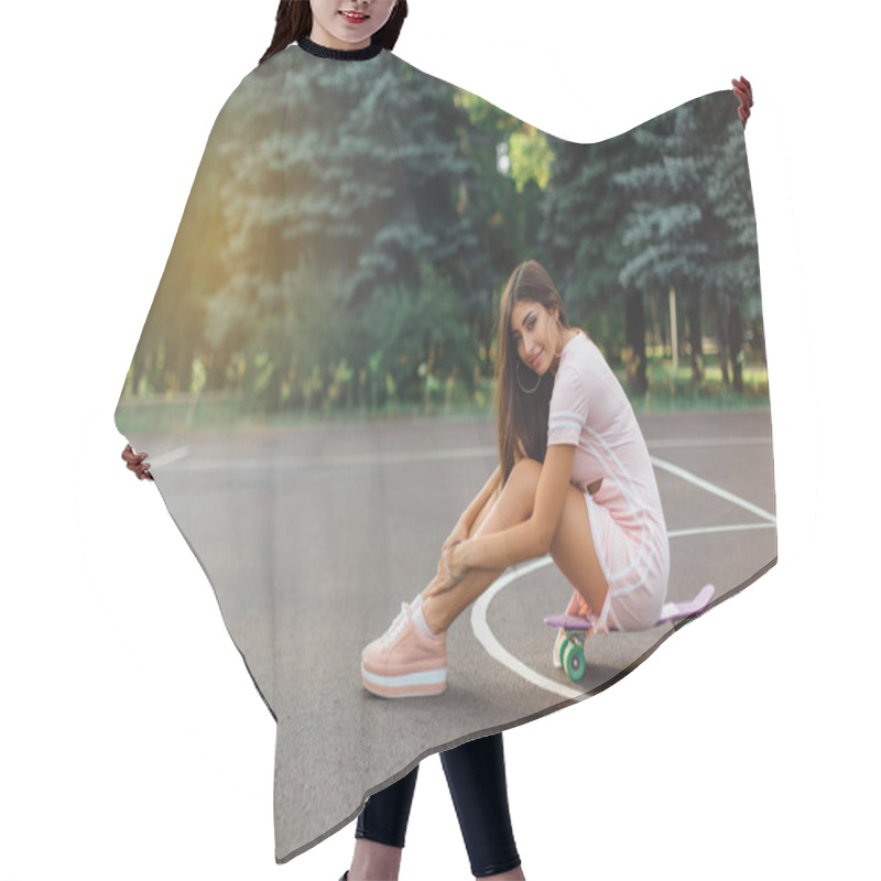 Personality  Portrait Of A Smiling Charming Brunette Female Sitting On Her Skateboard On A Basketball Court. Happy Woman With Trendy Look Taking Break During Sunset. Hair Cutting Cape