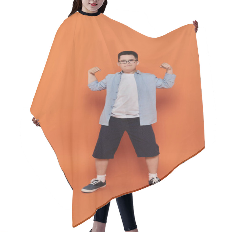 Personality  A Young Boy With Down Syndrome Flexes His Muscles In Front Of An Orange Background. Hair Cutting Cape