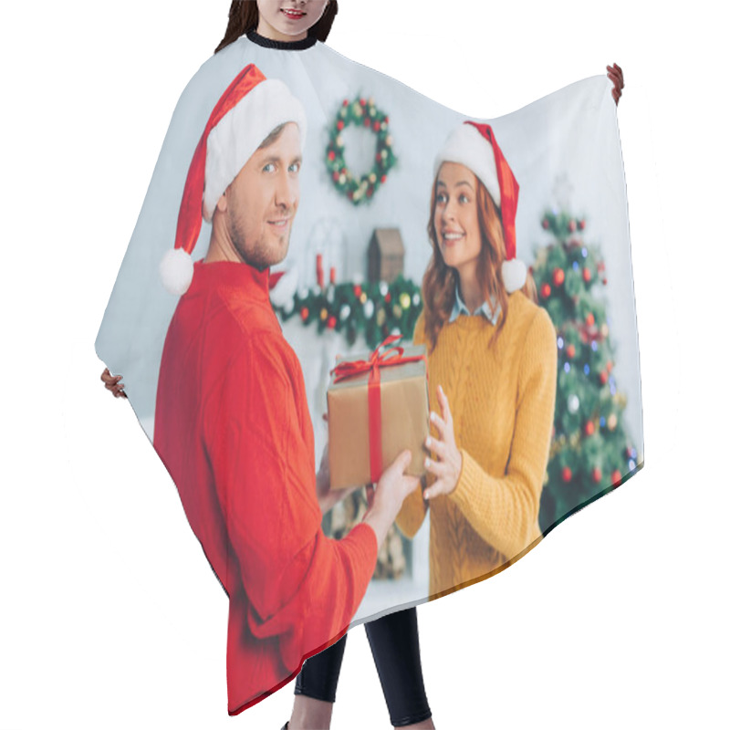 Personality  Smiling Man In Santa Hat Looking At Camera While Presenting Gift Box To Excited Wife Hair Cutting Cape