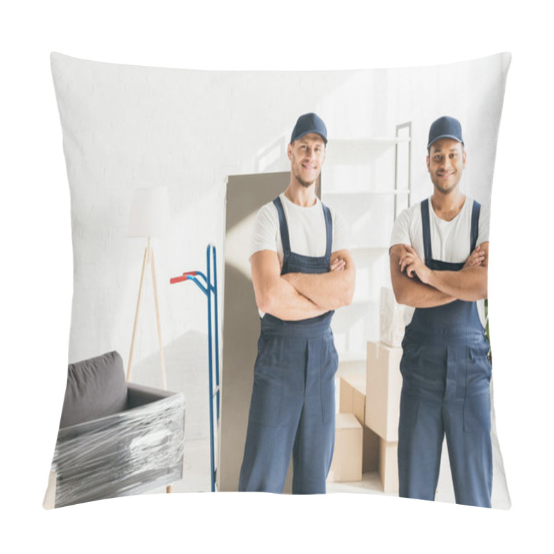 Personality  Cheerful Multicultural Movers In Caps Standing With Crossed Arms Near Wrapped Furniture  Pillow Covers