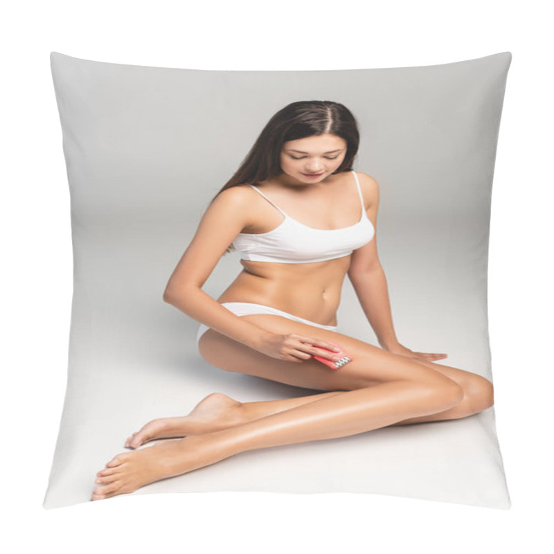 Personality  Young Adult Woman In White Underwear Shaving Leg With Pink Epilator, While Sitting On Grey Pillow Covers