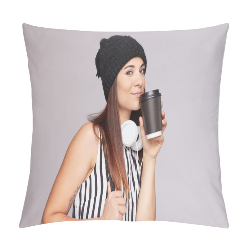 Personality  Studio Image Of Positive Young Woman In Knitted Hat, Casual Clothes, Smiling Flirty At Camera, Has Headphones On Neck, Holds Disposable Cup With Cappuccino, Carries Backpack, Isolated On Grey Wall. Pillow Covers