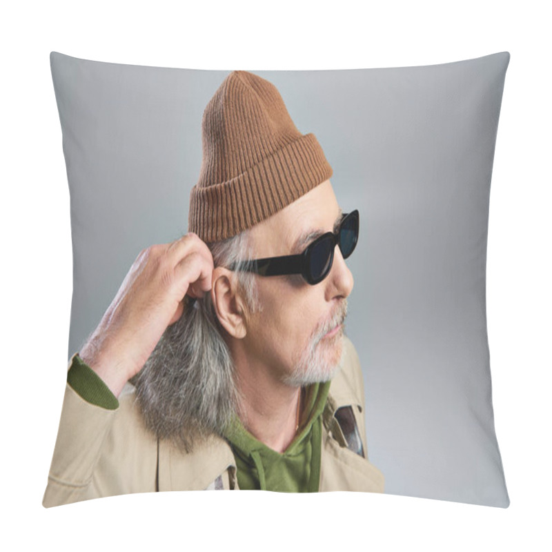 Personality  Portrait Of Elderly And Bearded Hipster Style Senior Man In Dark Sunglasses, Beanie Hat And Beige Trench Coat Adjusting Grey Hair And Looking Away On Grey Background, Fashionable Aging Concept Pillow Covers