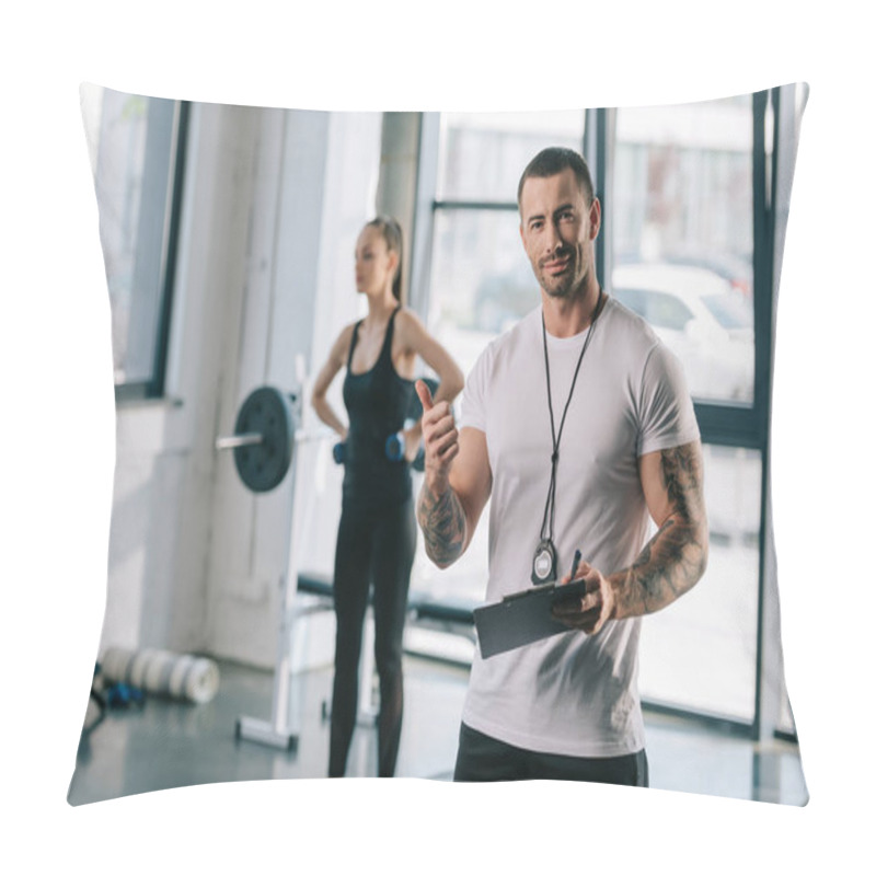 Personality  Personal Trainer With Clipboard Doing Thumb Up Gesture And Young Sportswoman Exercising With Dumbbells Behind At Gym Pillow Covers