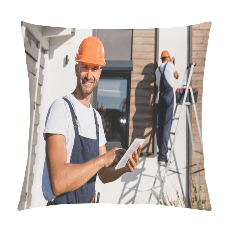 Personality  Selective Focus Of Builder With Digital Tablet Looking At Camera While Colleague Working On Ladder Near House At Background  Pillow Covers