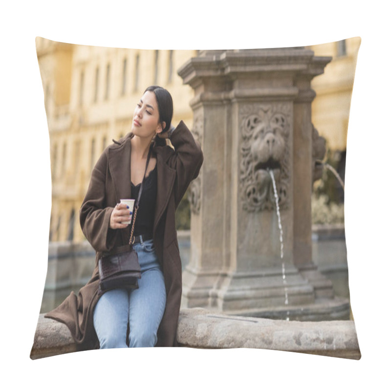 Personality  Young woman in coat touching hair and holding paper cup near blurred fountain in Charles Square in Prague  pillow covers