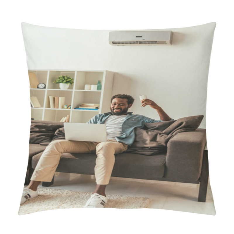 Personality  Cheerful African American Man Using Laptop And Holding Air Conditioner Remote Controller While Sitting On Sofa At Home Pillow Covers