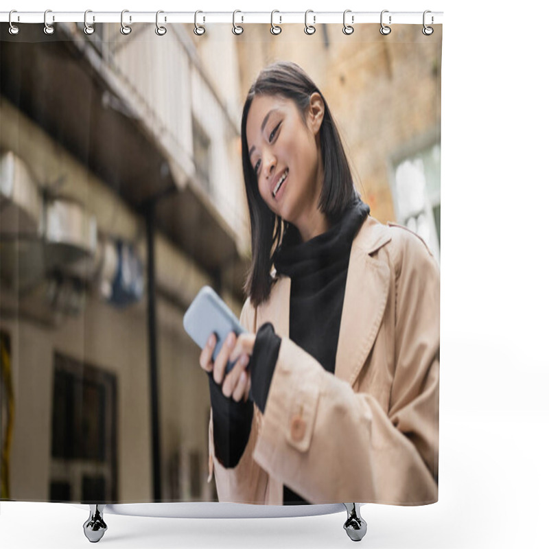 Personality  Low Angle View Of Smiling Asian Woman In Trench Coat Using Smartphone Outdoors  Shower Curtains