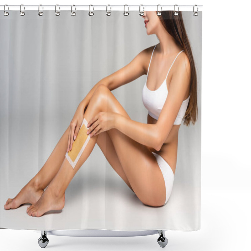 Personality  Cropped view of young adult woman in white underwear applying wax stripe on leg on grey shower curtains